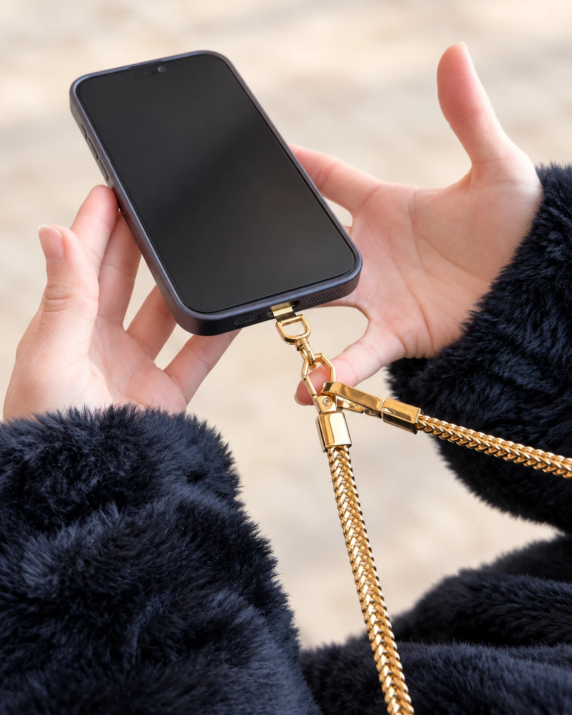 Manos sujetando un móvil con funda; el conector universal sale por la abertura inferior de la funda y la cadena AMARA AURA dorada está enganchada a él